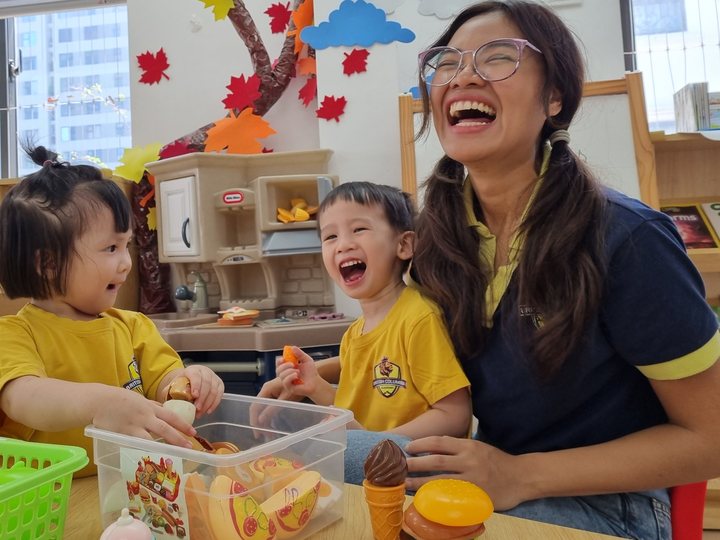 Toddlers laughing with their teacher during a role-play cooking activity